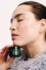 a brunette girl holding mini moisturizer that's green with red nails and a blue fur coat