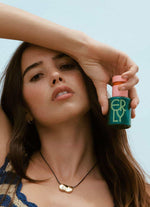 girl with brown hair and necklace holding green and pink skincare bottle